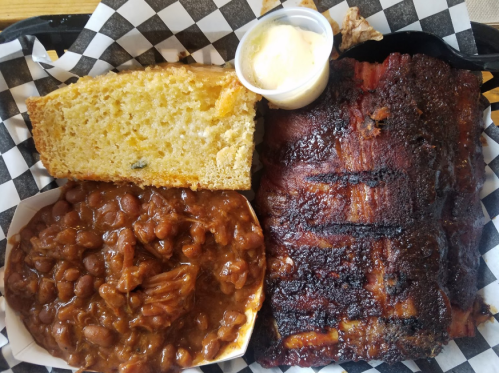 A plate featuring BBQ ribs, baked beans, cornbread, and a small cup of butter.