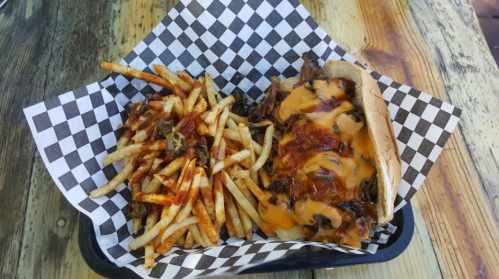 A plate with a cheesy beef sandwich and a side of seasoned fries on a checkered paper.
