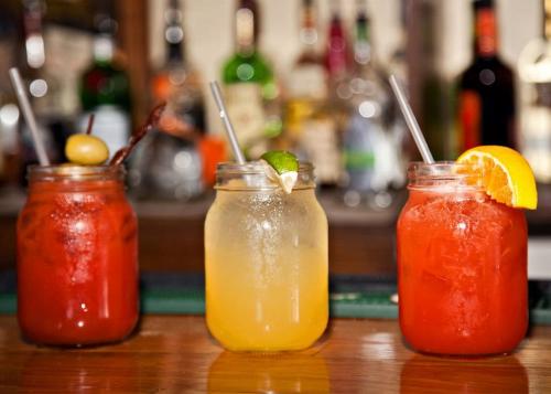 Three colorful cocktails in mason jars on a bar, with a blurred background of liquor bottles.