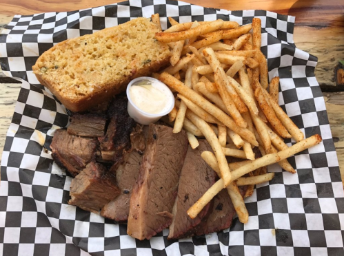A plate of sliced brisket, golden fries, and a piece of cornbread, served with a small cup of sauce on a checkered paper.