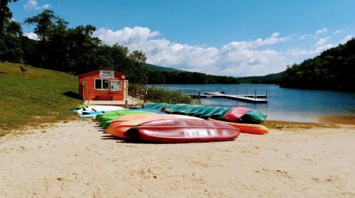 Colorful kayaks lined up on a sandy beach near a lake, with a boat rental shack in the background and blue skies above.
