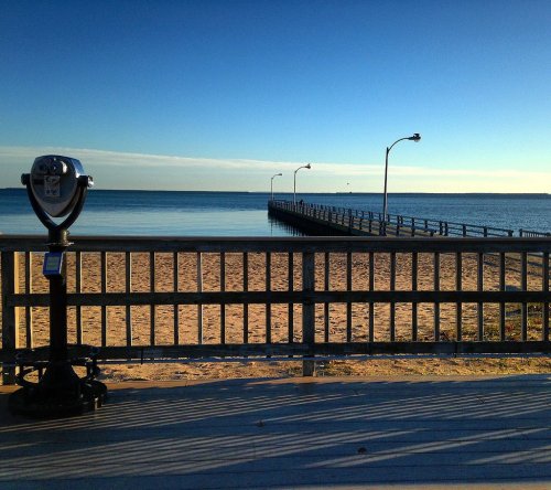 A beach view with a coin-operated viewer, a wooden pier extending into calm waters, and a clear blue sky.