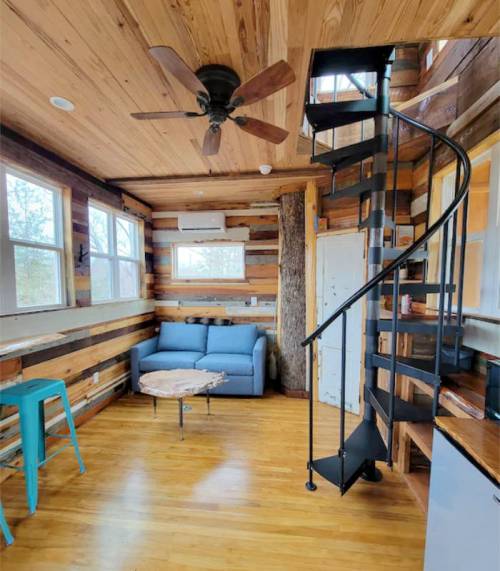 Cozy interior of a wooden cabin featuring a spiral staircase, blue couch, and large windows for natural light.