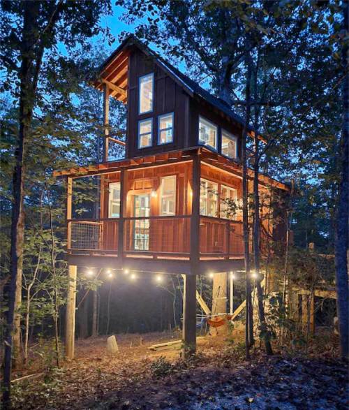 A modern treehouse illuminated at dusk, surrounded by trees, with large windows and a cozy, inviting design.