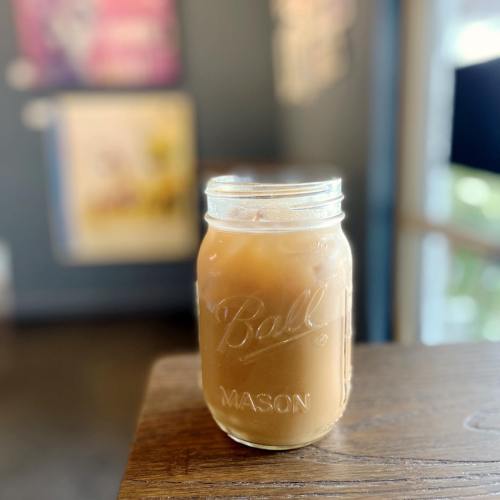 A glass mason jar filled with iced coffee sits on a wooden table, with a blurred background of colorful artwork.