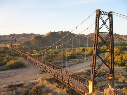 A suspension bridge spans a river, surrounded by rugged hills and desert vegetation under a clear sky.
