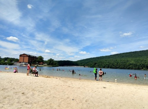 A sandy beach with people swimming and relaxing by a lake, surrounded by green hills and a blue sky with clouds.