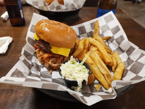 A hearty burger with cheese, pulled pork, and fries, served on a checkered paper plate with a side of coleslaw.