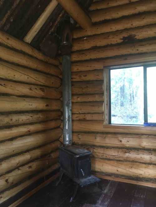 Interior of a rustic log cabin featuring a metal stove and a window with a view of the outdoors.