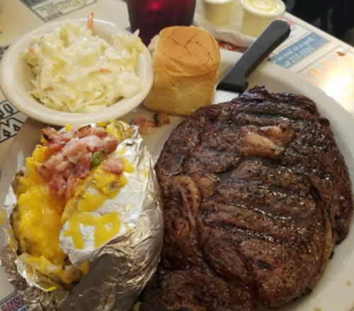 A plate featuring a large steak, loaded baked potato, coleslaw, and a roll, served in a casual dining setting.