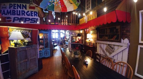 A cozy, colorful dining area with string lights, a long table, and vintage decor, featuring a sign for "Penn Hamburger."