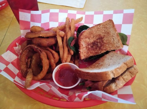 A red basket with a sandwich, onion rings, French fries, and a small cup of dipping sauce on a checkered paper.