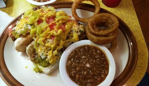 A plate featuring a loaded hot dog topped with mustard, onions, and relish, alongside onion rings and a bowl of baked beans.