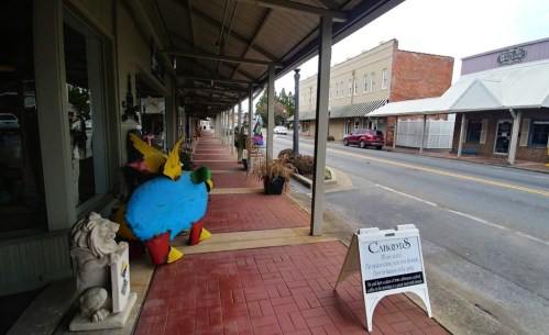 Colorful sculptures line a quaint sidewalk, with shops and a street visible in the background.