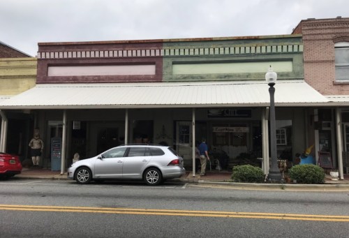A street view of a small town with colorful storefronts, a parked car, and a person browsing outside a shop.