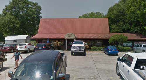 A restaurant with a red metal roof, surrounded by parked cars and greenery, on a sunny day.