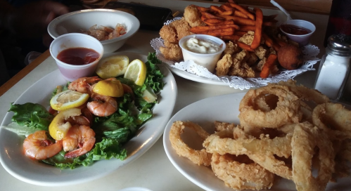 A table with two plates: one with shrimp salad and lemon, the other with fried chicken, sweet potato fries, and onion rings.