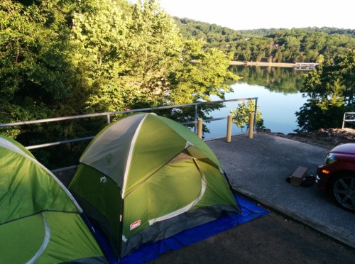 A green tent set up by a calm lake, surrounded by trees and reflecting sunlight.