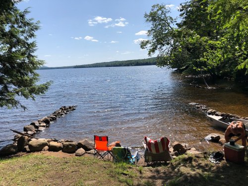 A serene lakeside scene with two chairs, a rocky shoreline, and a canoe by the water under a clear blue sky.