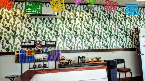 Colorful papel picado decorations hang above a patterned wall, with a counter displaying various items and a shelf of bags.