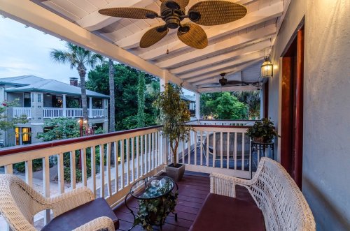 A cozy balcony with wicker furniture, a glass table, and a ceiling fan, surrounded by greenery and a peaceful atmosphere.