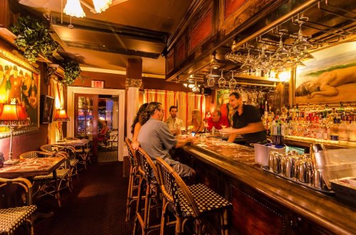 A cozy bar scene with patrons seated at the counter, a bartender serving drinks, and warm lighting throughout.