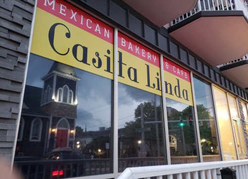Sign for "Casita Linda" Mexican Bakery & Café, reflecting a nearby building in the window.