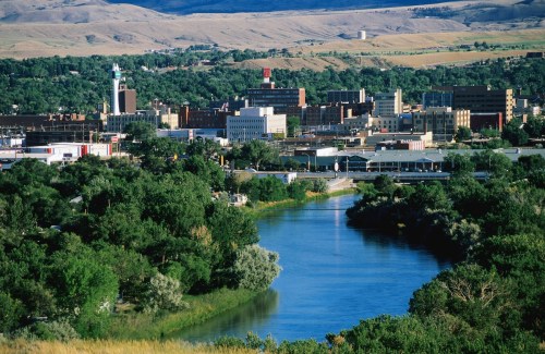 A scenic view of a city by a river, surrounded by greenery and hills in the background.
