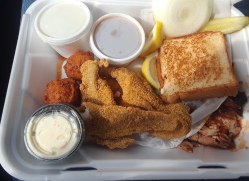 A takeout container with fried fish, toast, onion, lemon, dipping sauces, and hushpuppies.