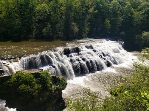 A serene waterfall cascades over rocks, surrounded by lush green trees and a calm river below.