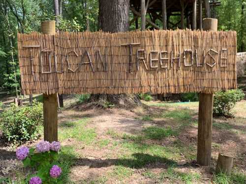 A wooden sign reading "Tucan Treehouse" stands in a garden with green foliage and purple flowers nearby.