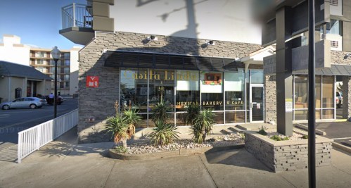 Exterior of a Mexican bakery and café with a stone facade, plants, and large windows. Nearby parking visible.