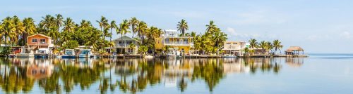 Colorful houses and palm trees line a calm waterfront, reflecting in the clear blue water under a sunny sky.