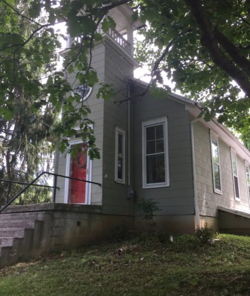A small, light green house with a bell tower, surrounded by trees and grass, featuring a red door and stone steps.