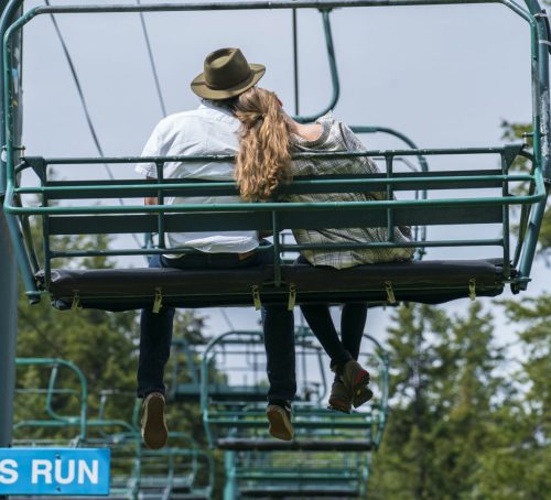 A couple sits closely on a ski lift, enjoying a moment together against a backdrop of trees.