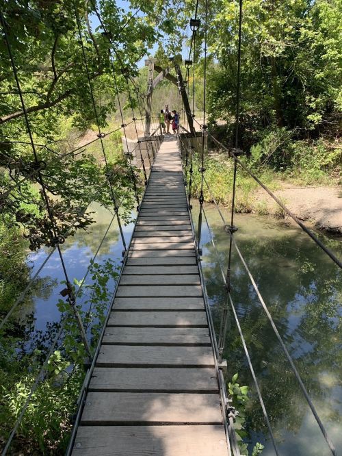 A wooden suspension bridge over a calm stream, surrounded by lush greenery and trees. People are walking in the distance.