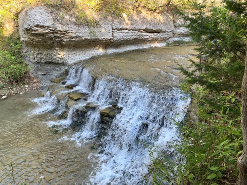 A serene waterfall cascading over rocky steps into a clear stream, surrounded by lush greenery and rocky cliffs.