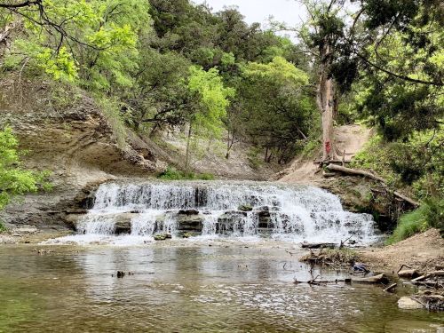 A serene waterfall cascades over rocks into a calm stream, surrounded by lush greenery and trees.