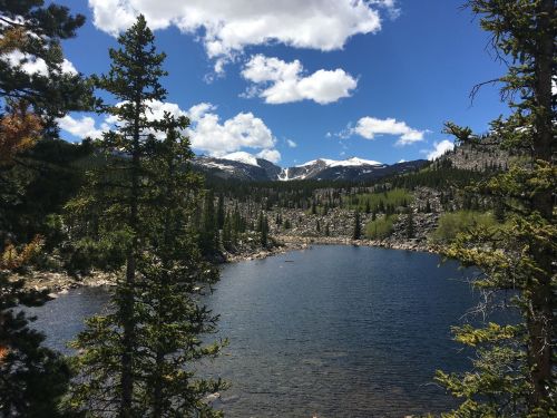 A serene lake surrounded by lush trees and mountains under a bright blue sky with fluffy clouds.