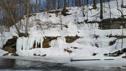 A snowy landscape featuring a rocky cliff with icicles and a frozen river below, surrounded by bare trees.