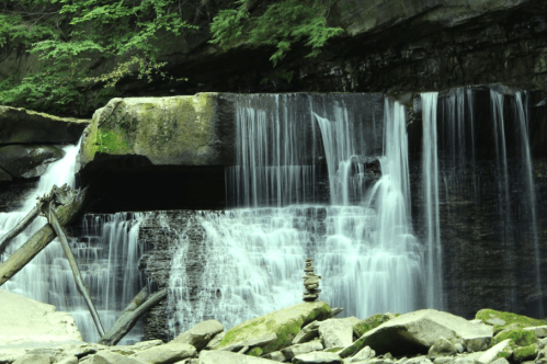 A serene waterfall cascades over rocks, with a stone tower balanced in the foreground amidst lush greenery.