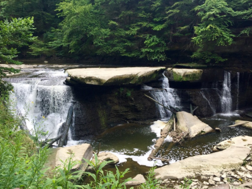 A serene waterfall cascading over rocky ledges, surrounded by lush green trees and a calm pool below.