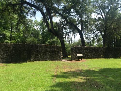 Ruins of a stone wall surrounded by lush greenery and trees, with a signpost in the foreground.