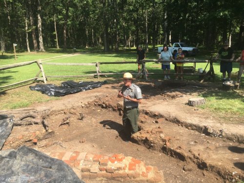 A park ranger leads a group at an archaeological dig site surrounded by trees and equipment.