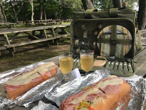 A picnic setup with two large sandwiches, two glasses of drink, and a picnic basket in a park with wooden tables.