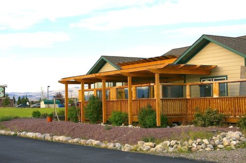 A wooden porch with a pergola extends from a light-colored house, surrounded by landscaped rocks and greenery.