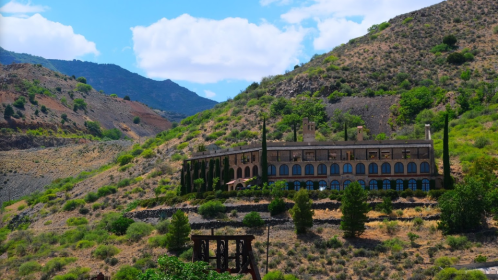 A large, abandoned building on a hillside, surrounded by greenery and mountains under a blue sky with clouds.