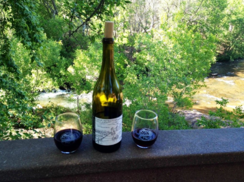 A bottle of wine with two glasses sits on a stone ledge, surrounded by lush greenery and a flowing stream in the background.