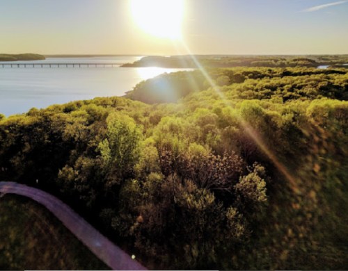 Aerial view of a sunlit landscape with a river, bridge, and lush green trees under a clear sky.