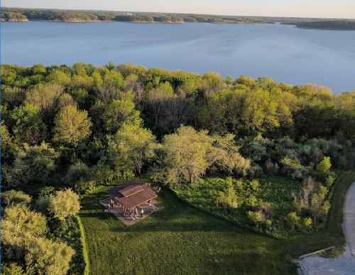 Aerial view of a lakeside cabin surrounded by lush greenery and calm water under a clear sky.